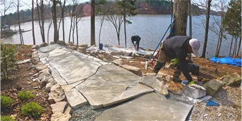 Workers installing a large stone pathway as part of a hardscaping project in Charlotte, NC, by the lakeside, showcasing precision and care in natural setting.